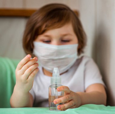 Küçük bir kız, tıbbi maskeli, elinde dezenfektan olan, COVID-19 salgını yüzünden karantinada oturuyor.