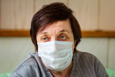 Yetişkin bir kadın, emekli bir kadın, tıbbi bir maske takıyor, dezenfektan tutuyor, COVID-19 salgını yüzünden karantinada oturuyor.