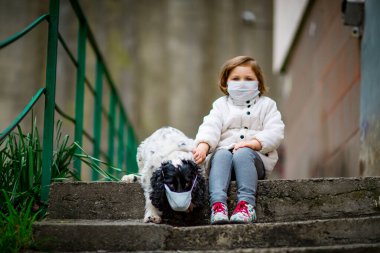 Tıbbi maskeli küçük bir kız covid-19 salgını sırasında bir köpekle bahçede yürür.