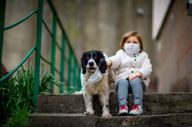 Tıbbi maskeli küçük bir kız covid-19 salgını sırasında bir köpekle bahçede yürür.