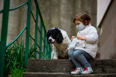 Tıbbi maskeli küçük bir kız covid-19 salgını sırasında bir köpekle bahçede yürür.