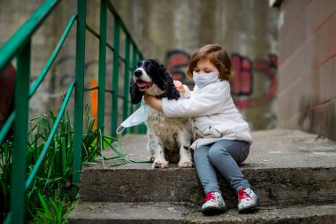 Tıbbi maskeli küçük bir kız covid-19 salgını sırasında bir köpekle bahçede yürür.