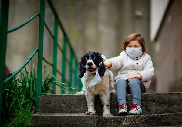 Tıbbi maskeli küçük bir kız covid-19 salgını sırasında bir köpekle bahçede yürür.