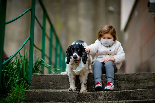 Tıbbi maskeli küçük bir kız covid-19 salgını sırasında bir köpekle bahçede yürür.