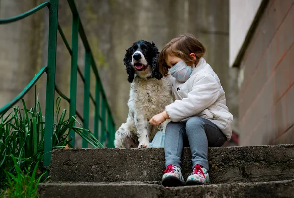 Tıbbi maskeli küçük bir kız covid-19 salgını sırasında bir köpekle bahçede yürür.