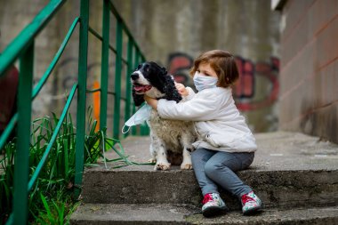 Tıbbi maskeli küçük bir kız covid-19 salgını sırasında bir köpekle bahçede yürür.