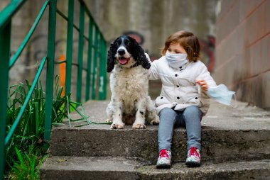 Tıbbi maskeli küçük bir kız covid-19 salgını sırasında bir köpekle bahçede yürür.