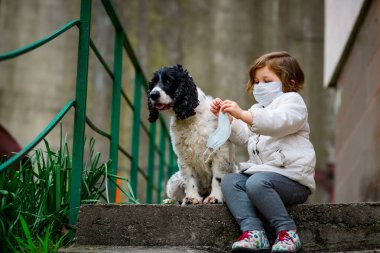 Tıbbi maskeli küçük bir kız covid-19 salgını sırasında bir köpekle bahçede yürür.