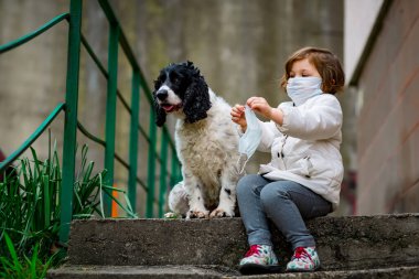 Tıbbi maskeli küçük bir kız covid-19 salgını sırasında bir köpekle bahçede yürür.