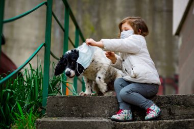 Tıbbi maskeli küçük bir kız covid-19 salgını sırasında bir köpekle bahçede yürür.