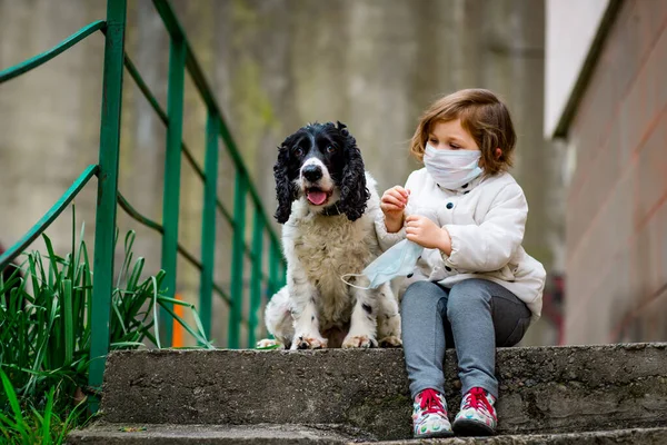 Tıbbi maskeli küçük bir kız covid-19 salgını sırasında bir köpekle bahçede yürür.