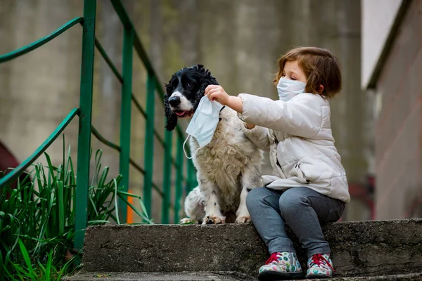 Tıbbi maskeli küçük bir kız covid-19 salgını sırasında bir köpekle bahçede yürür.