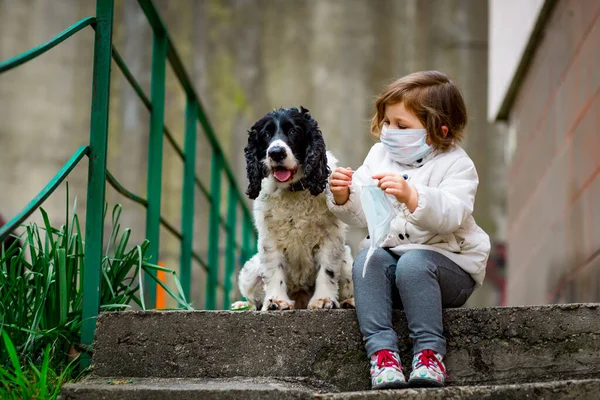 Tıbbi maskeli küçük bir kız covid-19 salgını sırasında bir köpekle bahçede yürür.