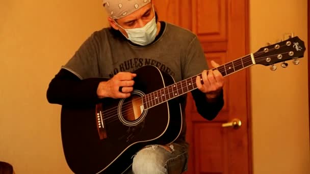 En man i medicinsk mask spelar gitarr, under karantän, på grund av covid-2019 pandemin — Stockvideo