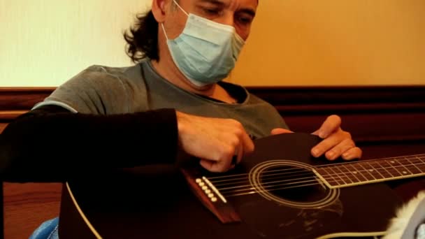 En man i medicinsk mask spelar gitarr, under karantän, på grund av covid-2019 pandemin — Stockvideo