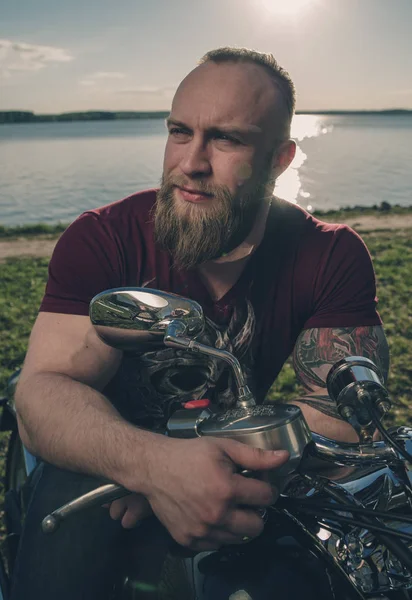 Biker on a motorcycle on the lake shore. Pensive romantic bearded biker in the setting sun. — Stock Photo, Image