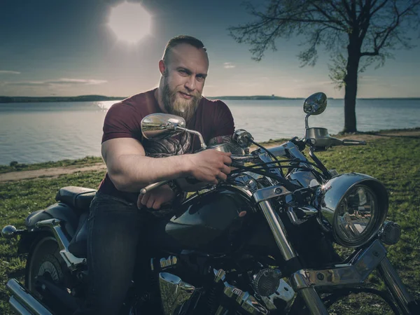 Motorkář na motorce na břehu jezera. Zamyšlený romantické vousatý biker v zapadajícím slunci. — Stock fotografie