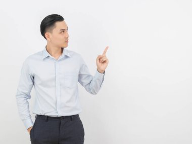 Young asian business man with blue shirt pointing to the side with a finger to present a product or an idea while looking forward smiling isolated on white background. clipart