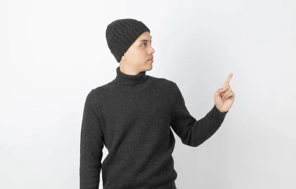 年轻英俊的亚洲男子 身穿灰色毛衣 头戴贝尼帽 用手指指向侧面 用白色背景来展示某种产品或想法 — 图库照片