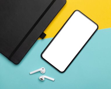 Beyaz ekran, kablosuz kulaklık ve sarı ve mavi arka planda izole edilmiş not defteri olan siyah akıllı telefon. Kesme yolu.