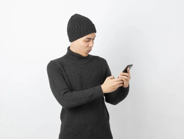 年轻英俊的亚洲男子 身穿灰色毛衣 头戴便帽 一边玩智能手机 一边带着白人背景的笑容 — 图库照片