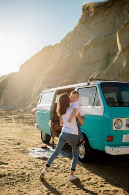 Genç esmer anne 3 yaşındaki oğluyla gün batımında bir retro otobüsünün yanında kumlu bir plajda oynuyor.