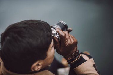 Kahverengi ceketli genç bir erkek fotoğrafçının, Avusturya, Plansee gölündeki ahşap bir göl evi güvertesinde küçük aynasız bir kamerayla fotoğraf çekerken yakın çekimi.