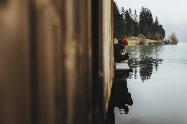 Göl planöründeki kayıkhane. Sonbahar havasında. Sis ve yağmur sırasında, kırmızı şapkalı kadın tahta güvertede oturuyor.