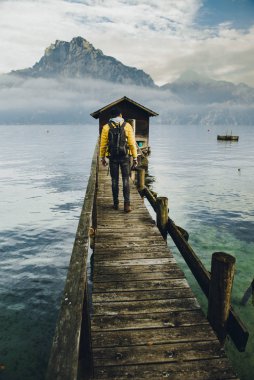 Sarı ceketli adam Traunsee Gölü 'ndeki tahta yaya köprüsünde yürüyor. Arka planda Traunstein tepesi alçak bulutlarla kaplı. Avusturya, Alp Bölgesi' nde yer alıyor..