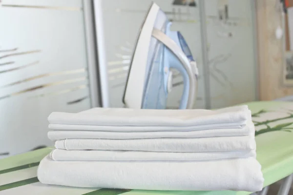 A stack of freshly washed white linen on an ironing board, an iron in the background. Clean clother, ironong concept. White fabric ironing after washing. Housekeeping