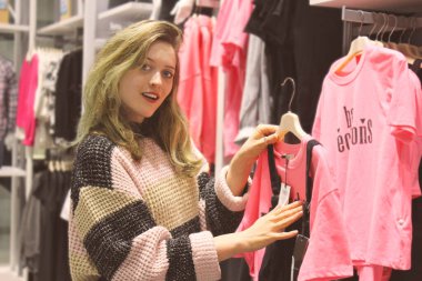 Elbise mağazasında alışveriş yaparken gülümseyen ve kameraya bakan güzel bir kadın. Yumuşak ışık, yumuşak renk arkaplan. Satış ve alışveriş konsepti.