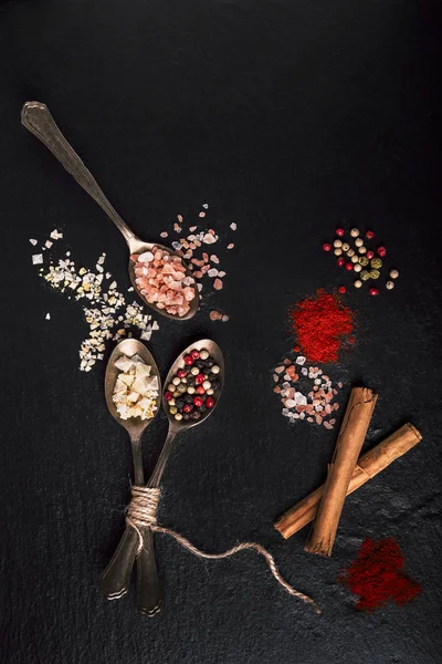 Spoons with flake salt, peppercorn and cinnamon on black stone