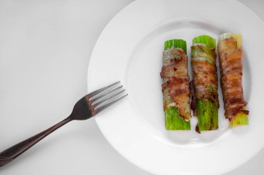 Pastırmaya sarılmış ve kızarmış üç kereviz sapı beyaz bir tabakta uzanıyor. Tabağın solunda bir çatal var.