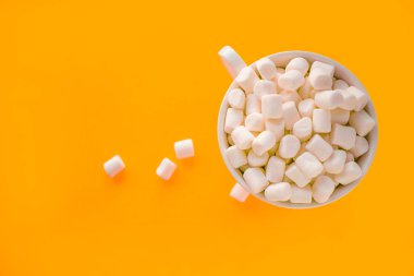 Sarı arka planda beyaz bir kupada küçük beyaz şekerlemeler. Kupa 'nın yanına birkaç marşmelov döküldü.