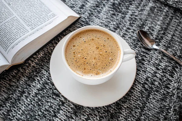 A white Cup of coffee on a gray background. Next to it is a teaspoon and an open book. The book quotes from the Bible