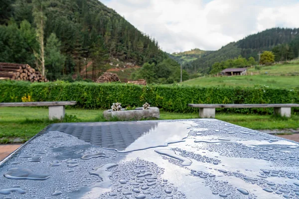 Raindrops on a metal table after the rain in the village with mountain, forest and piles of firewood at the background. Selective focus. — Stock Image Raindrops on a metal table after the rain in the village with mountain, forest and piles of firewood at the background. Selective focus. Stock Photo