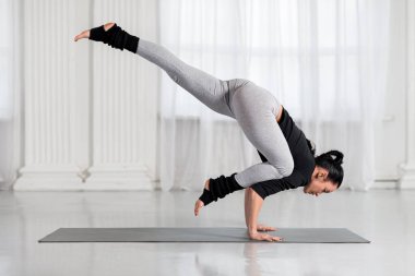 Asyalı bir kadın beyaz salonda çalışıyor, el ele tutuşup yoga yapıyor, tek bacaklı Crane Poz veriyor.,