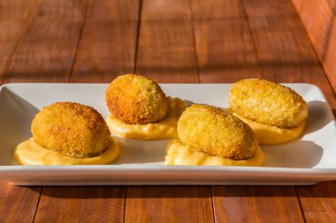 Geleneksel İspanyol atıştırmalığı yağda kızartılmış kroketler, beyaz tabakta sarı krema sosu, güneşli mutfakta. Ahşap arkaplanda yan görünüm.