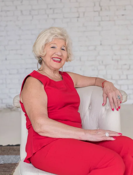 Modieuze oudere vrouw in een rode jurk zit in een witte stoel tegen een witte bakstenen muur glimlachend en poseren — Stockfoto
