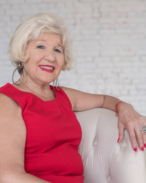Modieuze oudere vrouw in een rode jurk zit in een witte stoel tegen een witte bakstenen muur glimlachend en poseren — Stockfoto