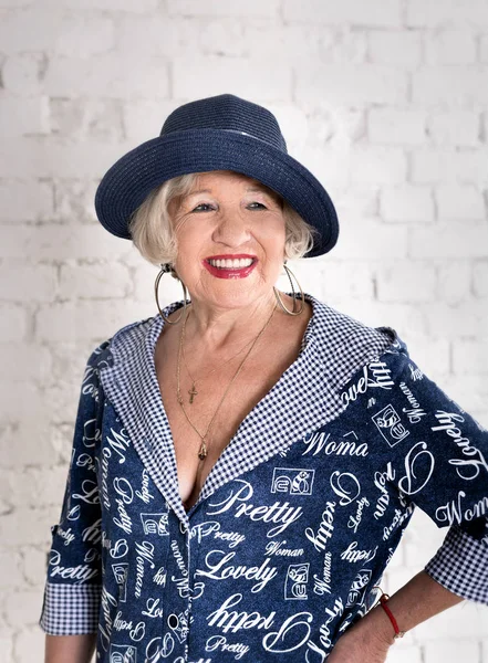 Fashionable elderly woman in a blue dress and hat against a white brick wall background smiling and posing — Stock Photo, Image