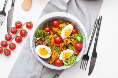 Kavrulmuş balkabaklı salata, haşlanmış yumurta, kiraz domatesleri, marul ve kabak tohumları gri kasede. Kara çatal ve ahşap tahta yakında. 