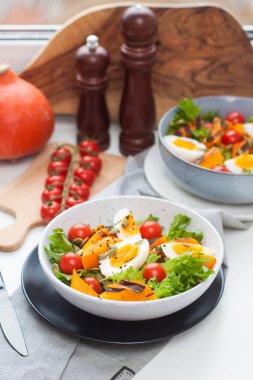 Kiraz domatesli yemek kasesi, kızarmış balkabağı, yeşil salata, tohum ve haşlanmış yumurta. Beyaz arka planda gri plaka. 