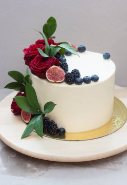 Taze kırmızı güllerle süslenmiş krem peynirli düğün pastası, incir ve yabanmersini ahşap pasta standında ve gri arka planda.. 