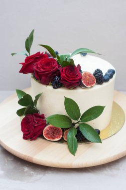 Taze kırmızı güllerle süslenmiş krem peynirli düğün pastası, incir ve yabanmersini ahşap pasta standında ve gri arka planda.. 