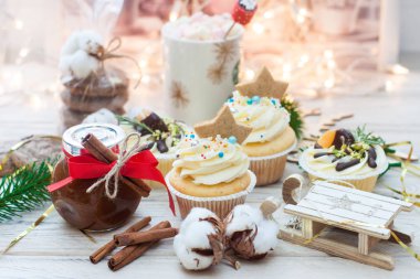 Krem peynirli kekler ve mandalinalar, pamuk çiçekleri, noel ağacı dalları, mini ahşap kızak, çikolatalı kurabiyeler, kırmızı riboonlu karamel kavanozu, ahşap zemin üzerinde tarçın ruloları.