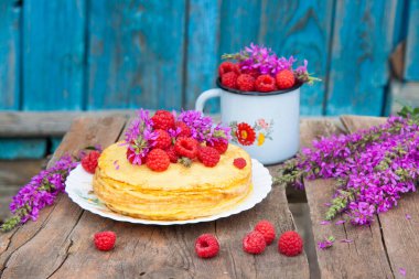 Pile of pancakes with fresh raspberries, old enamel bowl and field purple flowers on wooden table