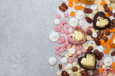 Çeşitli pembe, turuncu, kahverengi ve yeşil çikolatalı kalpler. Gri arka planda fındık ve çikolata damlaları var.