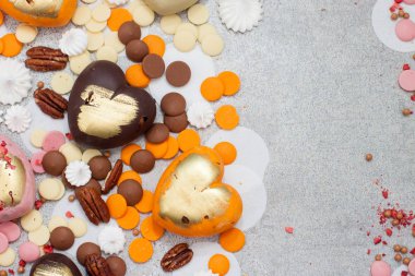 Çeşitli pembe, turuncu, kahverengi ve yeşil çikolatalı kalpler. Gri arka planda fındık ve çikolata damlaları var.