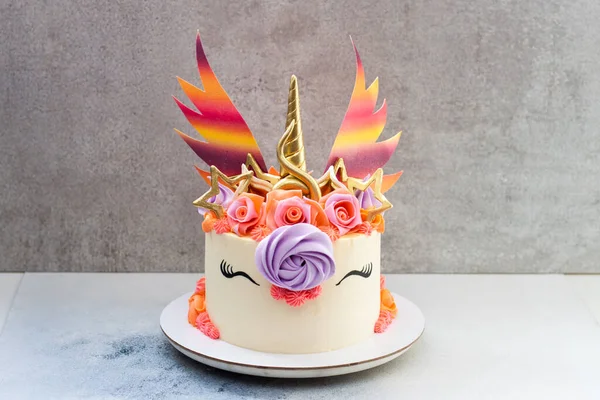 Bright and colorful unicorn cake with golden horn and ears, rainbow wings, roses and whipped cream decor. Grey background.